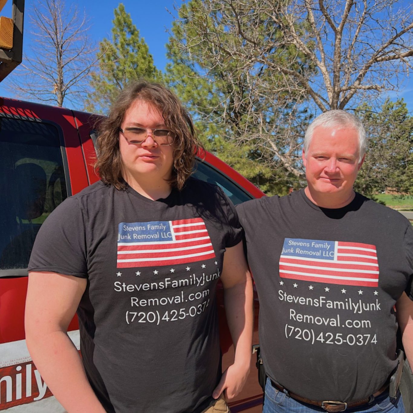 Two individuals wearing matching black shirts with an American flag design stand outdoors.