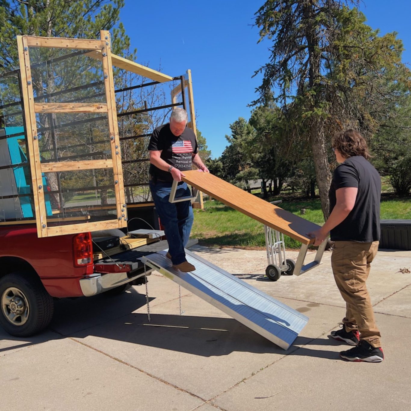 Two individuals unloading a large wooden plank from a truck ramp outdoors.